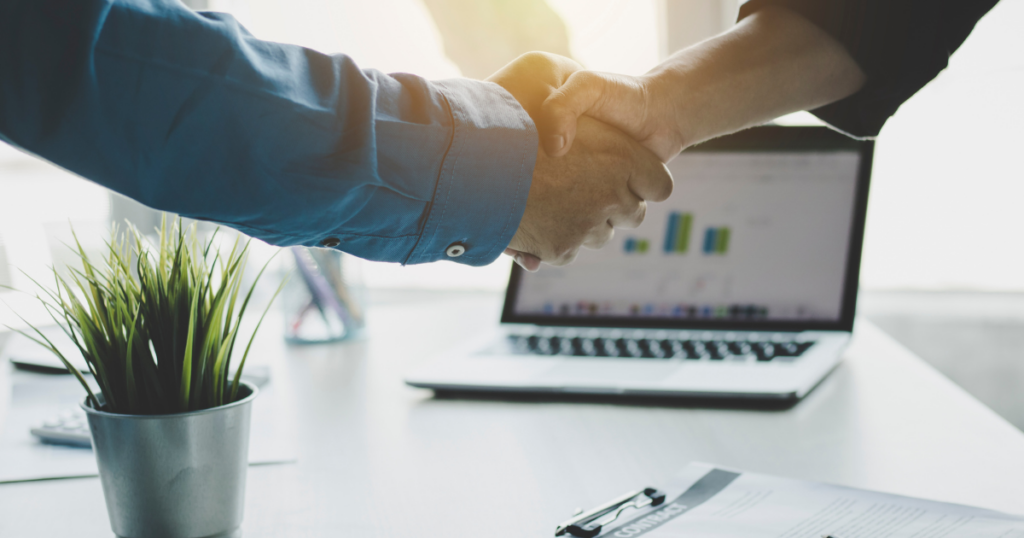 窓際のテーブルにグラフが映るノートPCと小さな観葉植物があり、手前で男性が握手している。