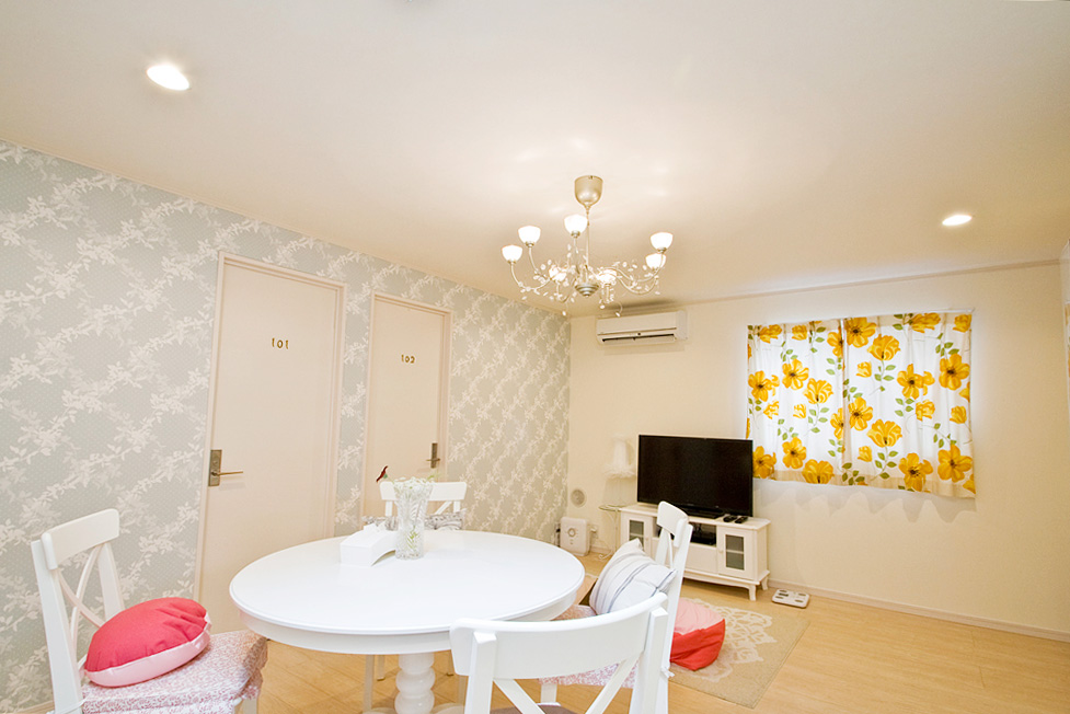 オレンジの光に包まれた上品な室内。シャンデリアとパンジー柄カーテン、白い家具と丸椅子にピンクのクッションが置かれている。