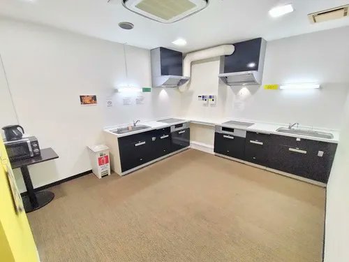 コーナーに収まった黒いピアノブラックのシンクと収納棚、レンジフードがあるおしゃれなキッチン。左奥には黒いレンジとミニポットが置かれ、手前のフロアは広々としてミルクティー色の床が広がっている。