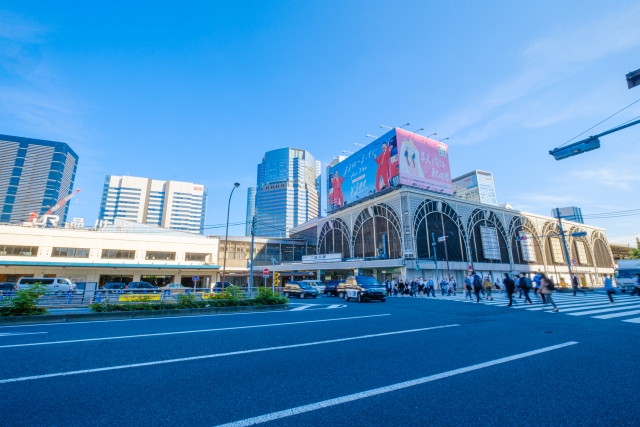 広い大きな道路に人々が颯爽と歩いている。横断歩道の向こうには看板広告が大きく掲げられたデパートのような商業施設が並び、都会的な賑わいを感じさせる街並み。