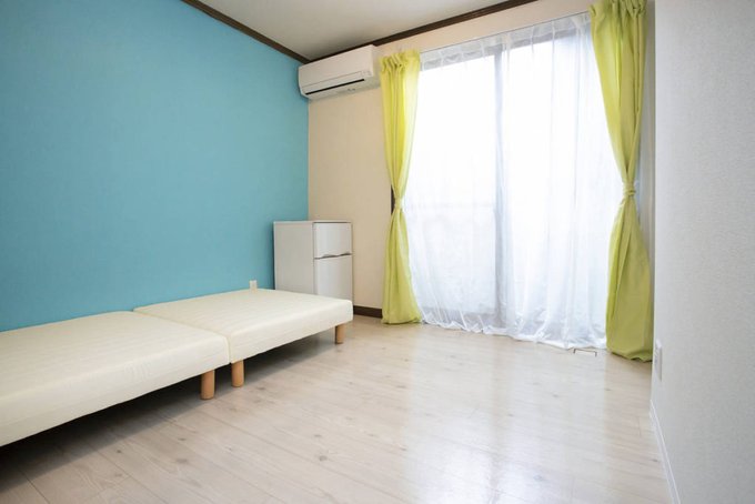 左側の壁一面がターコイズブルーで、低めの木の足付きアイボリーのソファが置かれている。大きな窓にはレースカーテンと両サイドに黄色のカーテンが整えられ、ベランダがありそうな雰囲気。奥のコーナーには小さなミニ冷蔵庫と白いエアコンがある。