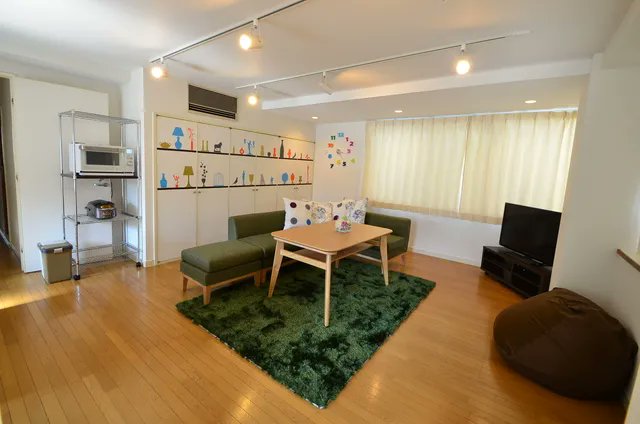 こげ茶のフローリングが広がる部屋。奥に深緑のソファがあり、白いサイコロ柄クッションが2つ置かれている。前には細長い白茶テーブルと真緑のラグマット。右奥の大きな窓には薄黄色のカーテンがかかり、壁面はカラフルなシールで飾られた北欧風の雰囲気。